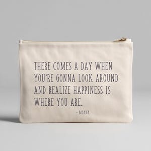 Könnte beinhalten: Eine cremefarbene Canvas-Tasche mit Reißverschluss. Auf der Tasche steht das Zitat "THERE COMES A DAY WHEN YOU'RE GONNA LOOK AROUND AND REALIZE HAPPINESS IS WHERE YOU ARE." in dunkelgrauem Text, zugeschrieben an Moana.