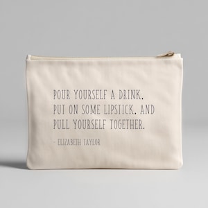 May include: Cream-colored zippered pouch with the quote "POUR YOURSELF A DRINK, PUT ON SOME LIPSTICK, AND PULL YOURSELF TOGETHER." attributed to Elizabeth Taylor. The pouch is rectangular and made of a soft fabric.