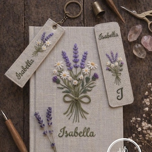 May include: Embroidered book and accessories featuring a lavender and daisy bouquet design. Includes a book, two bookmarks, and a keychain, all with the name "Isabella". The items are on a wooden surface with sewing tools.