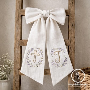 May include: Cream-colored linen sash with a large bow at the top. The sash features embroidered designs: a cross and the letter "T", each surrounded by a floral wreath. The sash is displayed on a wooden ladder.
