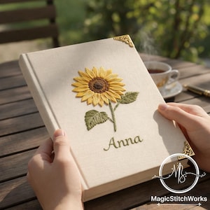Könnte beinhalten: Ein cremefarbenes Notizbuch mit einem detaillierten, gestickten Sonnenblumen-Design. Die Sonnenblume ist gelb und braun, mit grünen Blättern und Stiel. Der Name "Anna" ist darunter gestickt. Goldene Eckenschützer setzen Akzente.