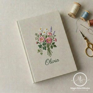 May include: A cream-colored journal with an embroidered floral design featuring pink roses, white daisies, and purple lavender. The name "Olivia" is embroidered below the flowers. Sewing supplies, including thread spools and scissors, are in the background.