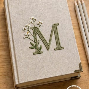 Könnte beinhalten: Beiges Notizbuch mit gesticktem grünem Buchstaben „M“ und floralem Design. Das Notizbuch hat goldene Eckenschützer und einen passenden Buchrücken. Drei Bleistifte sind rechts vom Notizbuch zu sehen.