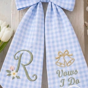 Puede incluir: Un lazo de cuadros azules y blancos con dos paneles colgantes. El panel izquierdo tiene una letra "R" verde bordada con detalles florales. El panel derecho presenta campanas doradas bordadas y el texto "Vows I Do".