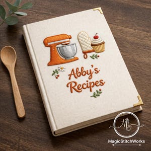 以下が含まれることがあります： クリーム色のレシピ本。金色のコーナープロテクターと、オレンジ色の刺繍で「Abby's Recipes」の文字。ミキサー、オーブンミトン、カップケーキの刺繍が施されています。木製のスプーンが木製の表面に置かれています。