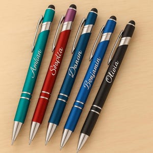 May include: Five personalized pens in teal, red, blue, navy, and black, each with a silver clip and tip. The pens are engraved with names: Aenlian, Soaplia, Danan, Benjamin, and Olivia. The pens are on a light wood surface.