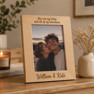 Op de afbeelding: Een lichtgekleurde houten fotolijst met de gegraveerde tekst "You are my today and all of my tomorrows" aan de bovenkant en "William & Kate" aan de onderkant, met een foto van een koppel dat buiten bij zonsondergang knuffelt.