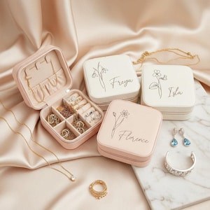 May include: A pink jewellery box, open to reveal rings and necklaces. Three square, personalised jewellery cases with names and floral designs are shown. Other jewellery, including earrings and a bracelet, is arranged nearby.