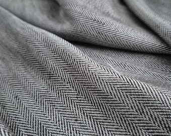 May include: Close-up of a grey herringbone fabric with a textured pattern. The fabric has a woven design with diagonal stripes, creating a V-shaped pattern. The fabric is folded, showing the texture and depth of the material.