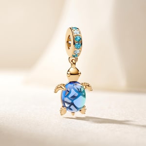 May include: A gold-toned charm featuring a sea turtle pendant with a translucent blue and white patterned shell. The bail is cylindrical and embellished with a row of alternating light blue and clear round stones set in gold-toned metal.