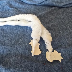 May include: Two white fuzzy worm toys with small black eyes and attached wooden feet rest on a blue fabric surface. The worms are made of a soft, plush material and are posed in a playful arrangement.