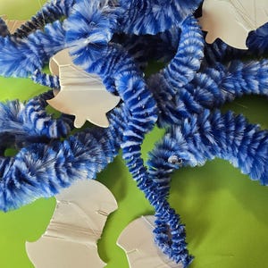 May include: Close-up of blue and white fuzzy garland with white bell-shaped cutouts. The garland is arranged in a circular pattern against a green background. The fuzzy texture and bright colors create a festive look.