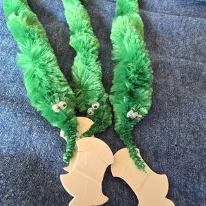 Op de afbeelding: Drie groene, fuzzy wormspeelgoed met wiebelogen en beige uitgesneden vormen. De wormen zijn gemaakt van groene pijpenragers en hebben kleine, ronde ogen. De beige vormen zijn aan de wormen bevestigd.