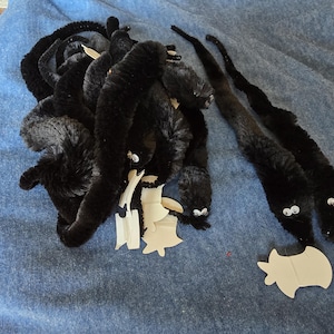 Puede incluir: Gusanos negros de limpiapipas con ojos saltones y recortes de cartón sobre una superficie azul. Los gusanos están dispuestos en un montón y dos están separados. Los recortes tienen forma de estrella.