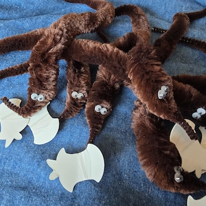 Pode incluir: Uma coleção de brinquedos de minhoca marrons e fofos com olhos arregalados e recortes de papel branco. Os brinquedos estão dispostos sobre uma superfície de tecido azul, criando uma cena lúdica. Estes brinquedos são para crianças.