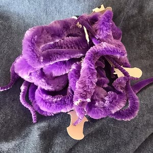 May include: A purple plush octopus toy with multiple tentacles. The toy has a soft, fuzzy texture and is on a blue denim surface. The octopus has white eyes and a tag attached.