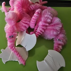 May include: A whimsical craft project featuring pink pipe cleaner worms with googly eyes, arranged with white paper cutouts on a green surface. The worms are fluffy and textured, creating a playful and imaginative scene.