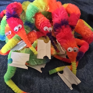 Puede incluir: Un conjunto de coloridos y peludos juguetes de gusanos en tonos rojos, naranjas, amarillos, verdes y azules. Cada gusano tiene ojos saltones y va acompañado de una pequeña etiqueta blanca. Los juguetes están colocados sobre una superficie de mezclilla azul.