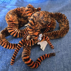 Può includere: Un giocattolo a forma di ragno fatto a mano, peloso, con strisce arancioni e nere, simile a una tigre. Il ragno ha gli occhi a palla ed è fatto di scovolini. Il giocattolo è appoggiato su una superficie di jeans blu.