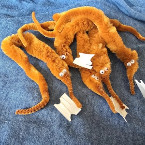 May include: Several orange fuzzy worm toys with googly eyes and white paper tags are arranged on a blue fabric surface. The worms are made of a soft, plush material and have a playful appearance.