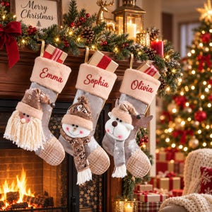 May include: Three silver sequined Christmas stockings with beige cuffs embroidered with the names Emma, Sophia, and Olivia. Each stocking features a plush character: a Santa Claus, a snowman, and a reindeer, with small gift boxes tucked inside.