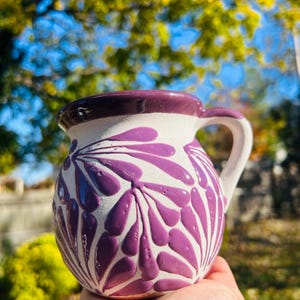 Könnte beinhalten: Eine handgefertigte Keramiktasse mit weißem Grund und einem lila Blumenmuster. Der Rand und der Henkel sind ebenfalls lila. Die Tasse wird in einer Hand vor einem verschwommenen Hintergrund aus grünem und gelbem Laub gehalten.