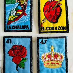 Peut inclure: Quatre écussons brodés avec des motifs colorés. L'un représente une femme dans un bateau, un autre un cœur percé d'une flèche, une rose rouge et une couronne dorée. Chaque écusson a du texte en espagnol.