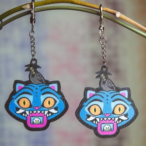 Może przedstawiać: Para niebieskich kolczyków w kształcie kota z pomarańczowymi oczami, białymi zębami i różowym językiem. Każdy kolczyk ma czarnego kruka w kapeluszu czarownicy. Kolczyki wiszą na srebrnych łańcuszkach.