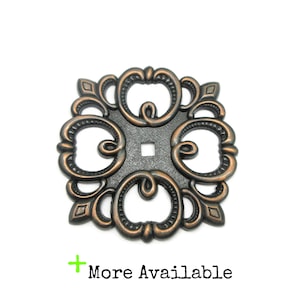 May include: A square-shaped metal furniture escutcheon plate with a dark bronze finish and copper-toned highlights. The piece features an intricate scrollwork pattern surrounding a central square hole. The text More Available is printed below.