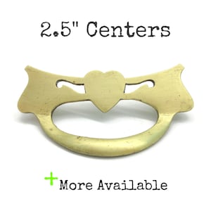 May include: A brass drawer pull with a central heart shape and curved arms, featuring the text 2.5" Centers and More Available. The metal hardware has a smooth, matte finish and a wide, arched handle design for furniture attachment.