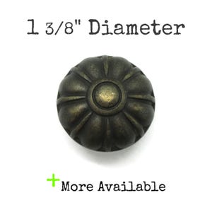 May include: A round, dark bronze-toned metal cabinet knob with a fluted, pumpkin-like design and a smooth center circle. The text above reads 1 3/8" Diameter and the text below reads + More Available, indicating the size and stock status.