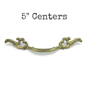 May include: Ornate brass-toned drawer pull with a curved design. The pull measures 5 inches (12.7 cm) between the centers of the mounting holes. The decorative hardware features scrolling leaf and floral details, and the text "5" Centers" is above the pull.