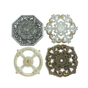 May include: Four decorative metal embellishments with intricate designs. The embellishments are in various shapes and sizes, including a round, square, octagon, and snowflake shape. The embellishments are in silver, gold, and white.