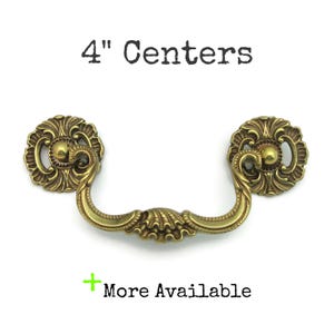 May include: A brass-colored drawer pull with ornate floral detailing. The pull has a curved handle and two decorative rosettes on either end. The text "4" Centers" is above the pull, and "More Available" is below.