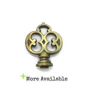 May include: A brass lamp finial featuring an ornate, openwork clover-like design with a central diamond shape and a tiered, circular base. The metal has a weathered, aged patina with visible dark highlights. The text More Available is present.