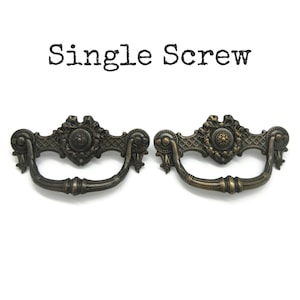 May include: Two antique bronze-colored metal drawer pulls with a swinging handle and ornate backplate featuring a central floral motif and lattice detailing. The text "Single Screw" is printed in black above the two hardware pieces.