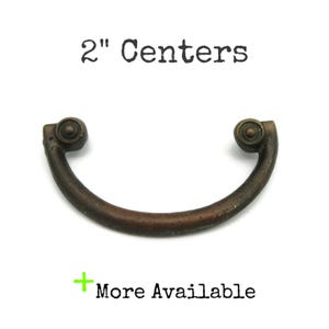 May include: A bronze-colored drawer pull with a curved handle and decorative end pieces. The text "2" Centers" is above the pull, and "More Available" is below. The pull measures 2 inches between the centers.