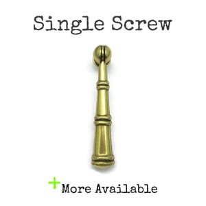 May include: A brass-toned metal drop pull handle featuring a vertical, tapered cylindrical body with horizontal ring detailing and a spherical top attachment. The text Single Screw is printed above the object, and + More Available is printed below.