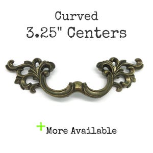 May include: A curved metal drawer pull with an antique brass finish and ornate scrollwork detailing. The text above the handle reads Curved 3.25" Centers, and the text below reads + More Available, indicating the specific mounting distance.