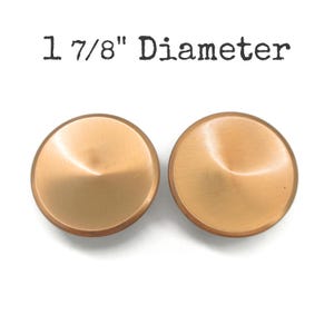 May include: Two copper-colored, round cabinet knobs with a diameter of 1 7/8 inches. The knobs have a brushed finish and a slightly domed shape. The text "1 7/8" Diameter" is printed above the knobs.