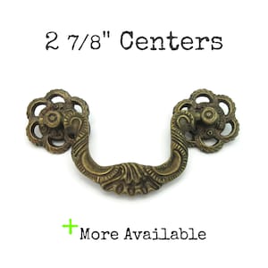 May include: Antique brass-toned drawer pull with floral accents. The pull has a curved handle and measures 2 7/8" between centers. The text "More Available" is also visible.