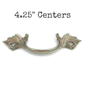 May include: A decorative cabinet pull with a curved handle and ornate leaf-like details. The pull is a muted silver color and measures 4.25 inches between the centers of the mounting holes. The text "4.25" Centers" is above the pull.