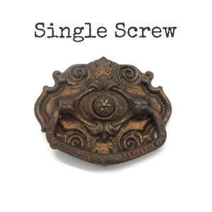 May include: A bronze-toned metal drawer pull with an ornate, symmetrical design featuring scrollwork and a central floral motif. A hinged, U-shaped handle is attached to the decorative backplate. The text "Single Screw" appears above the object.