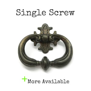May include: A dark bronze-toned metal drawer pull with a swinging oval handle and a decorative backplate. The text "Single Screw" appears above the hardware, and the text "+ More Available" is positioned below the object on a white background.