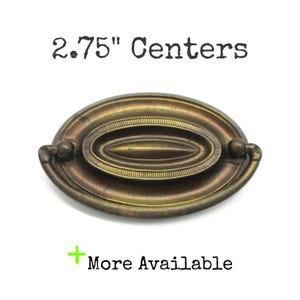 May include: An oval-shaped antique brass drawer pull with a central raised horizontal element and a swinging handle. The text 2.75" Centers is displayed above the hardware, and the text + More Available is displayed below the hardware.