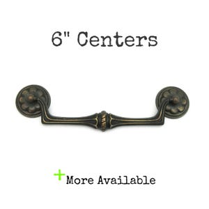 May include: Antique bronze-finished drawer pull with a floral design on the ends and a decorative center. The pull measures 6 inches (15.24 cm) between the centers of the mounting holes. The text "6" Centers" and "+ More Available" are visible.