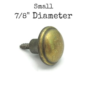 May include: A small, antique-style brass knob with a 7/8" diameter. The knob has a rounded, domed shape and a threaded screw for attachment. The text "Small" and "7/8" Diameter" is printed above the knob.