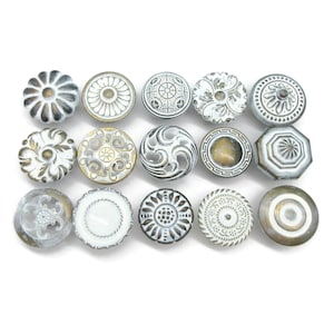 May include: A collection of fifteen round cabinet knobs arranged in three rows. Each knob features a distinct white and metallic gold or bronze finish with intricate embossed patterns, including floral, geometric, and swirling decorative designs.