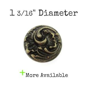 May include: A round, antique brass-colored button featuring a raised, swirling floral relief pattern. The text 1 3/16" Diameter is printed above the button, and the text + More Available is printed below the button on a white background.