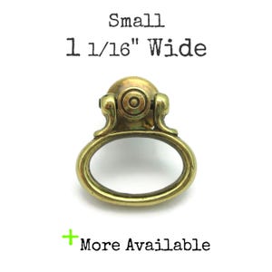 May include: Small, antique-style brass drawer pull with an oval handle and decorative backplate. The pull is 1 1/16" wide. The text "More Available" is visible.